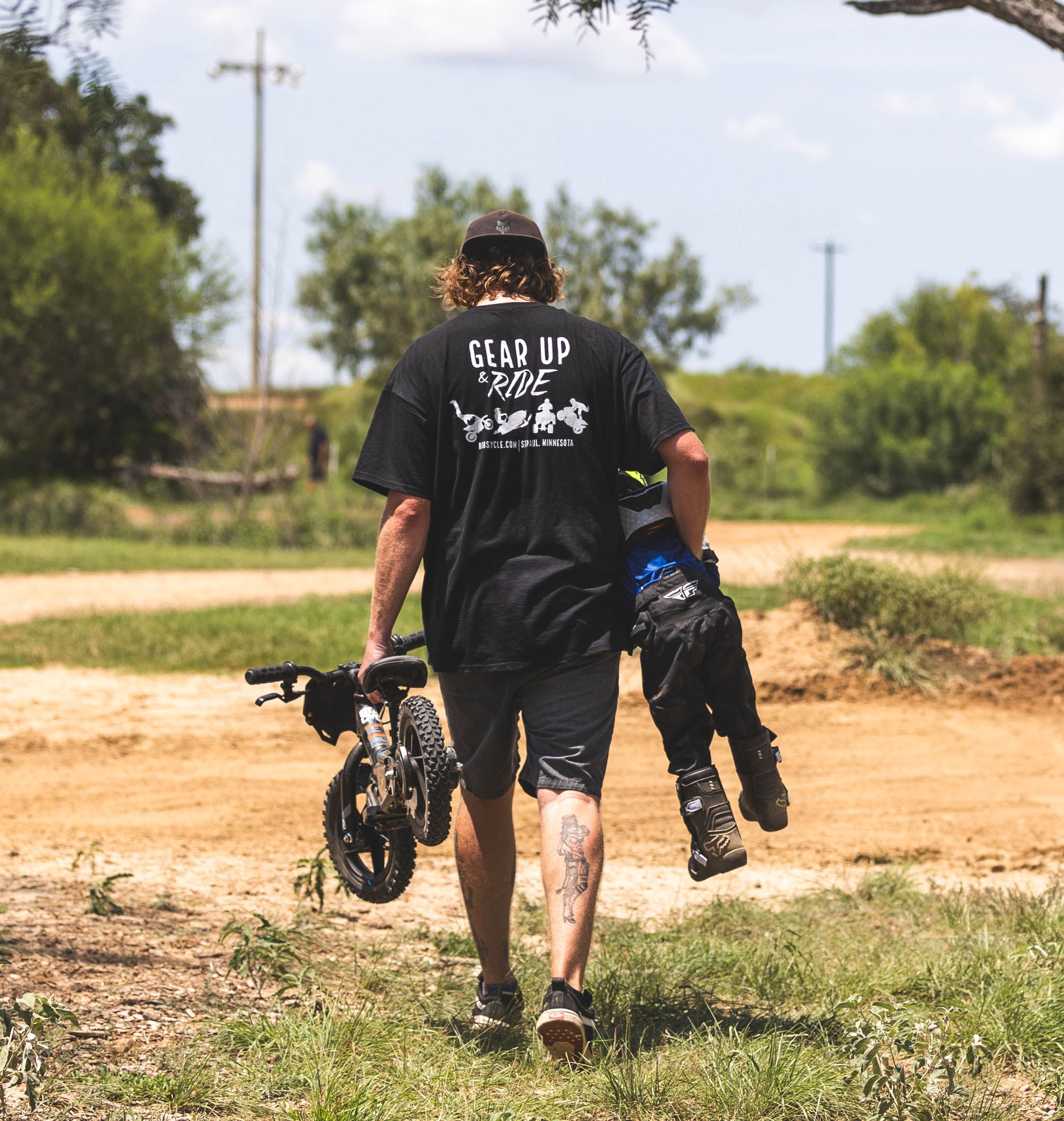 Rider walking with dirt bike gear and wearing a 'Live Up' t-shirt on dusty path.