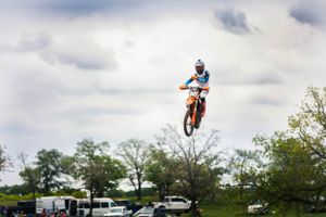 Dirt bike rider soaring high through the air during a motocross jump with trees and vehicles in the background.