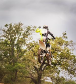 Motocross rider performing high-altitude jump on dirt track with autumn trees in background.