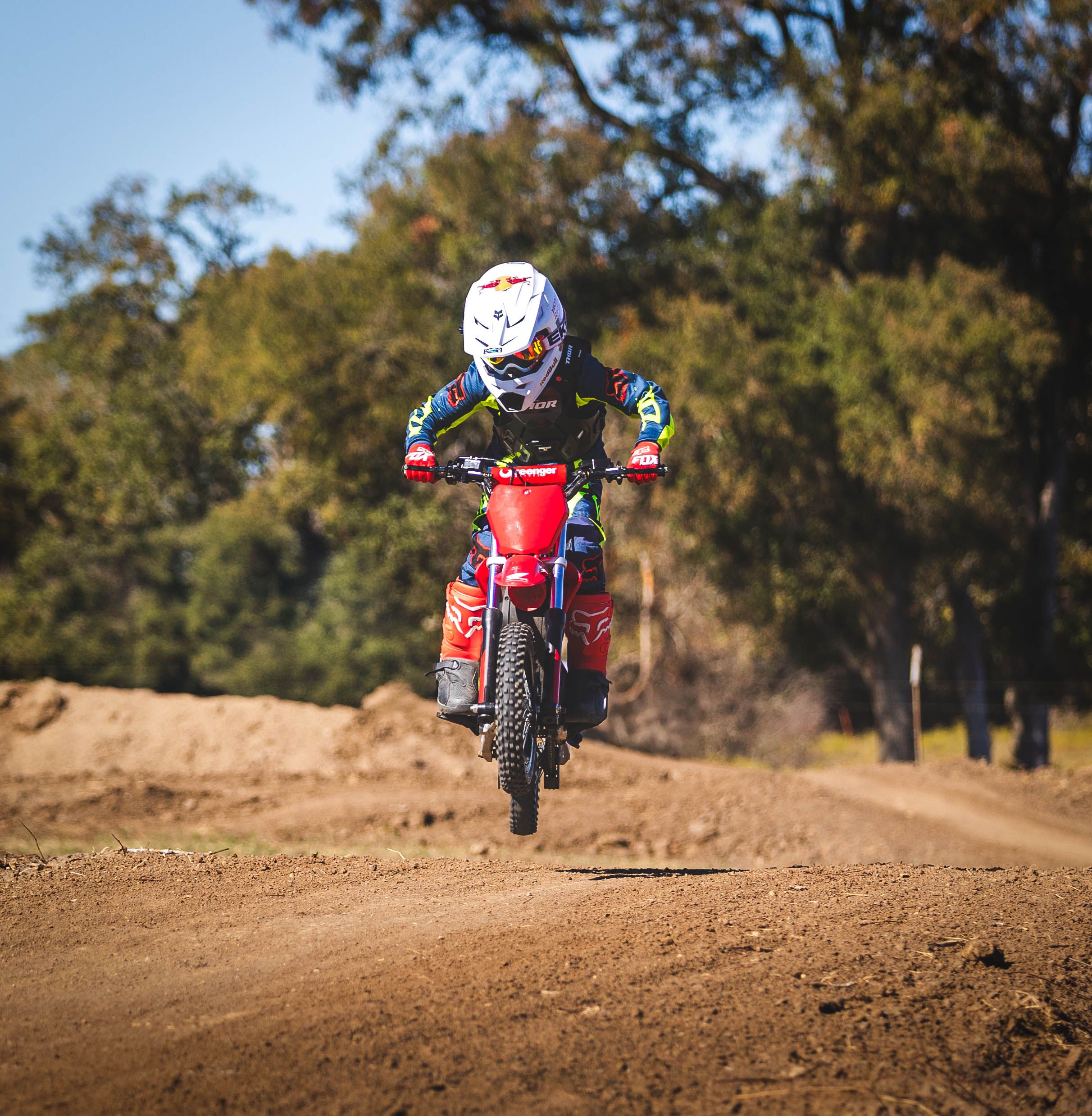 Motocross rider launching red dirt bike off jump on dusty track with trees in background.