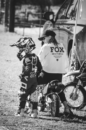Black and white photo of motocross gear and equipment by parked vehicle at dirt bike track.