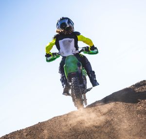 Motocross rider launching green dirt bike over jump against bright blue sky.