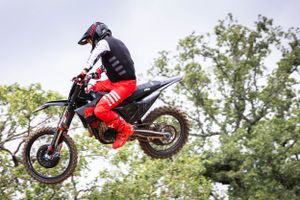 Motorcycle rider executing a high jump maneuver on a dirt bike against a cloudy sky.
