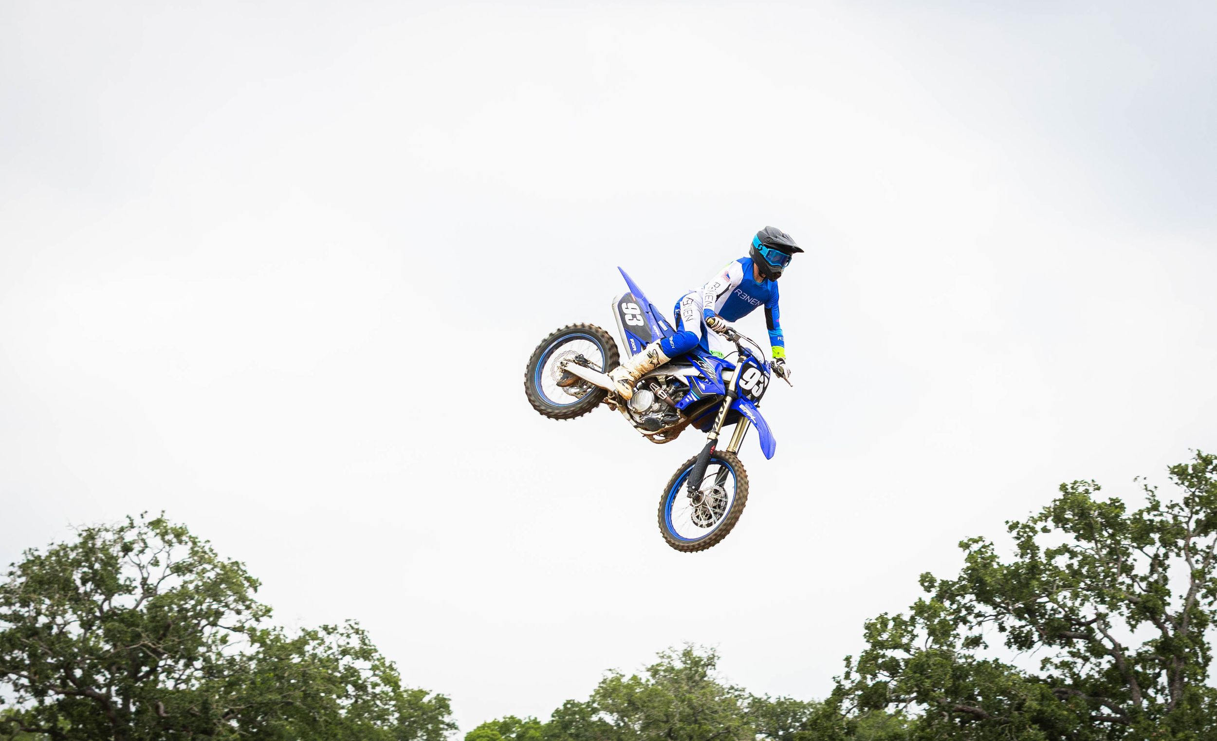 Dirt bike rider in blue gear performs aerial stunt with perfect form against bright sky.