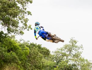 Dirt bike racer in blue gear performing sideways aerial maneuver against tree backdrop.