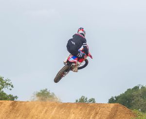 Dirt bike rider executing mid-air jump maneuver on red motorcycle over dirt track.