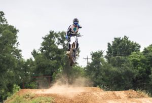 Motocross rider launching off dirt jump with trees in background.