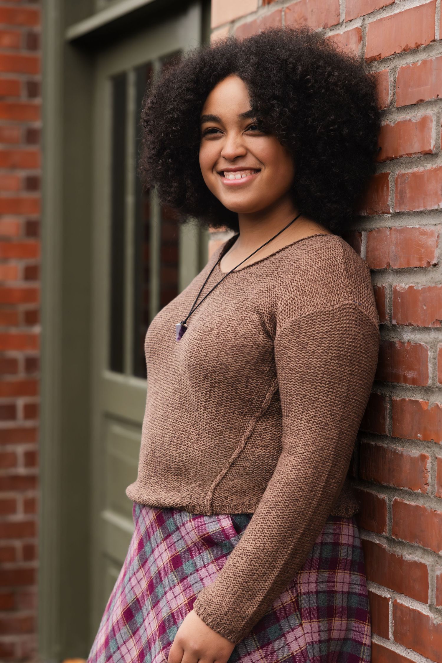 Woman in brown sweater and plaid skirt smiles while leaning against brick wall.