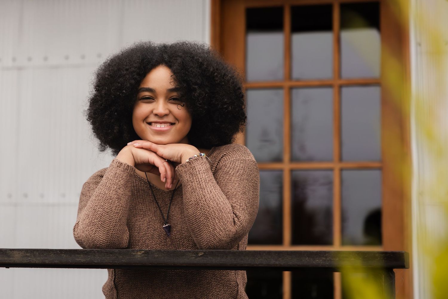 A person in a brown sweater leans on a railing while smiling outdoors.