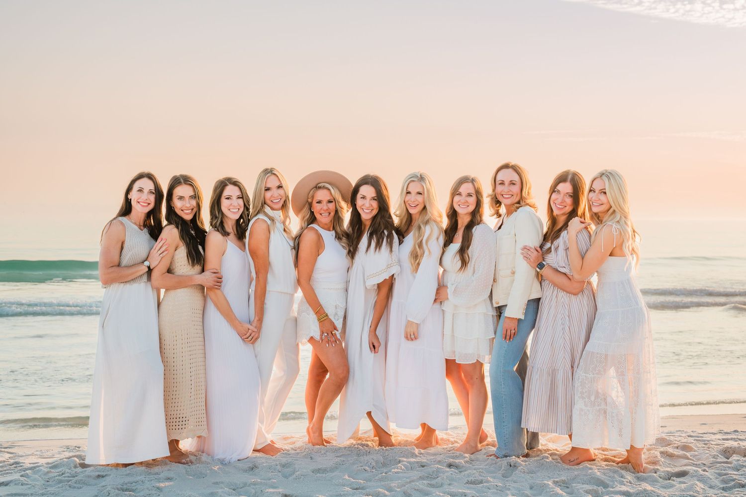 Group in white dresses and outfits standing together on beach at sunset with ocean waves in background.