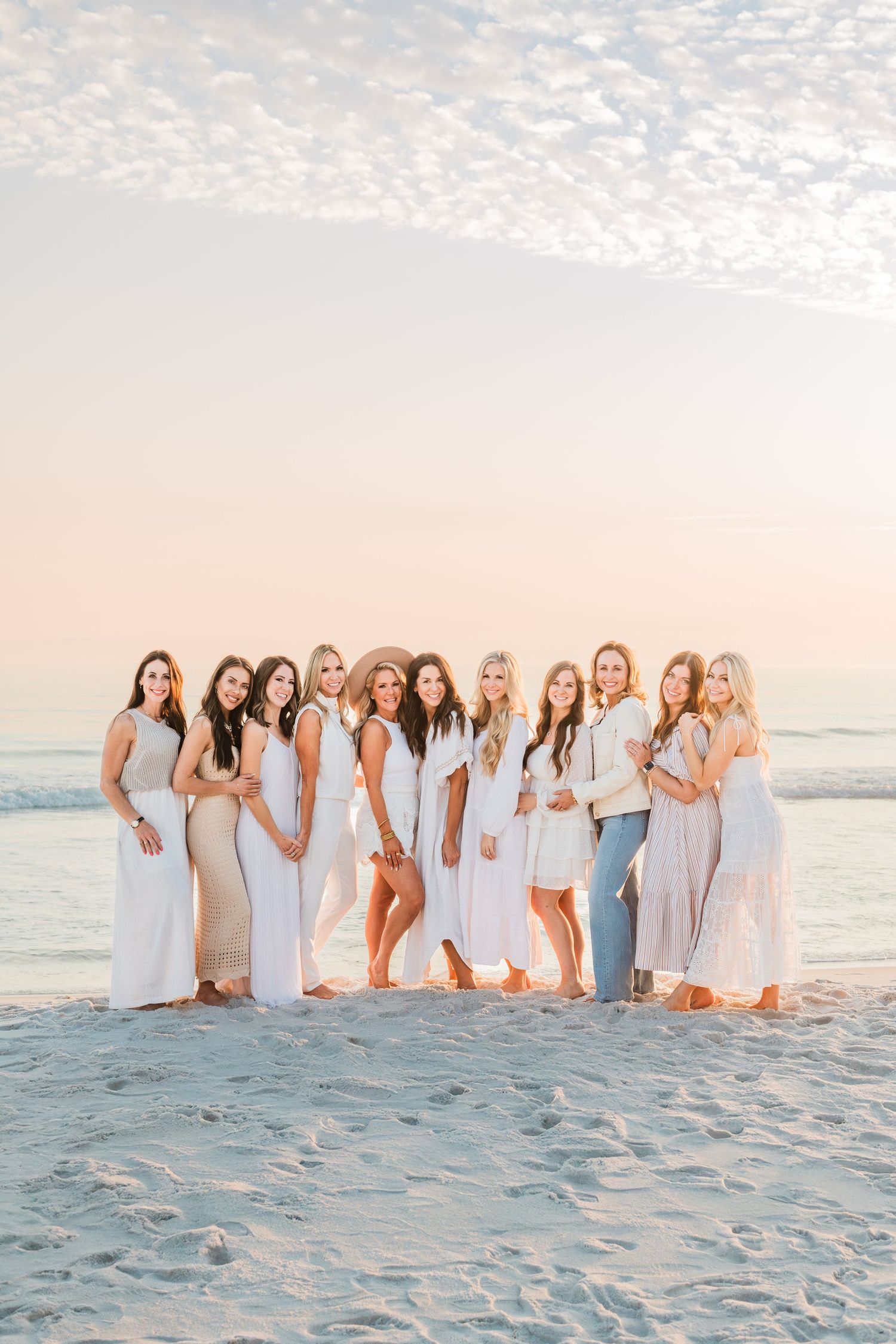 A group wearing coordinated white and light blue outfits poses together on a sandy beach at sunset.