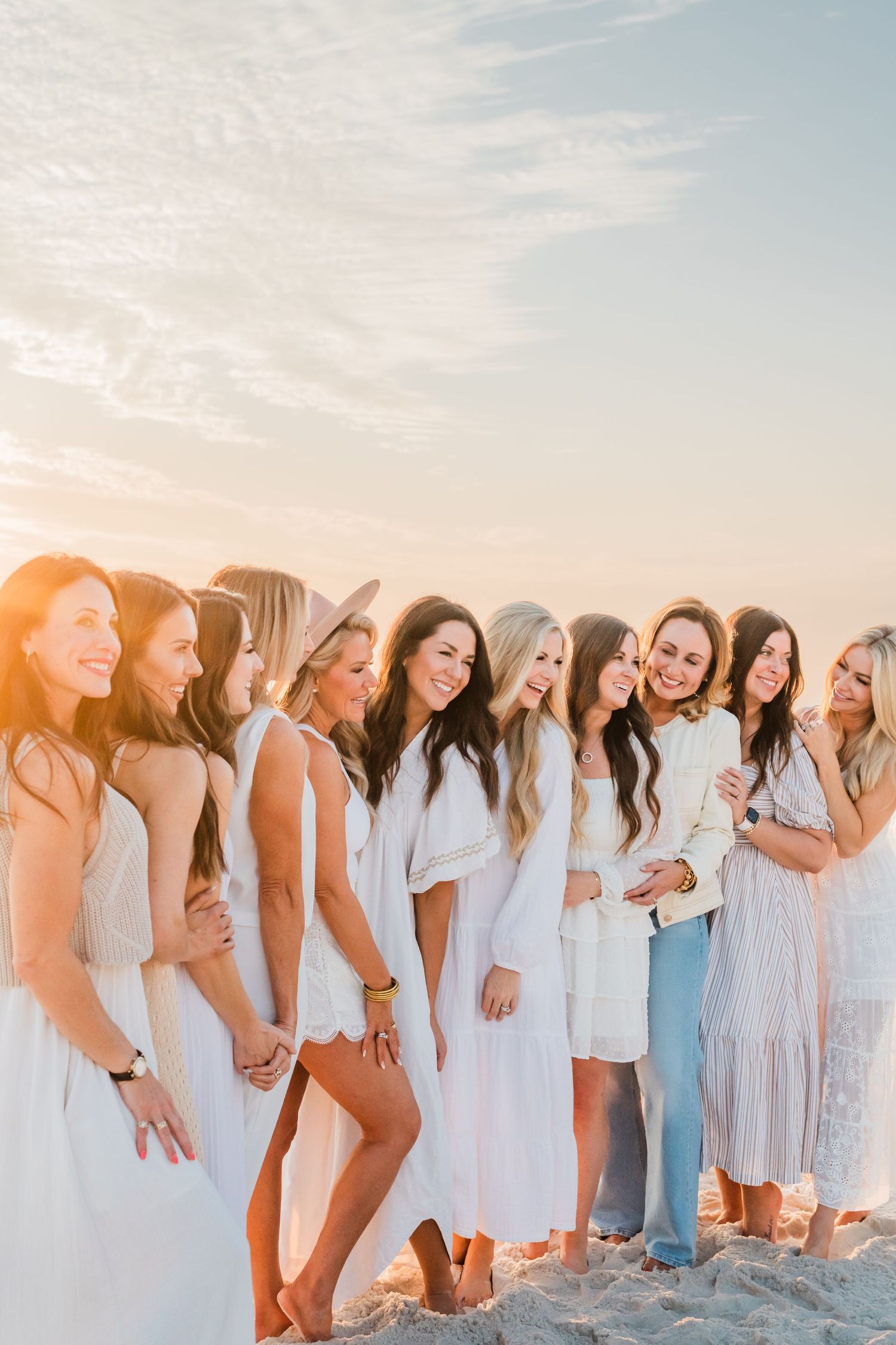 Group of friends in light colored dresses and outfits laughing together on a beach at sunset with golden sunlight.