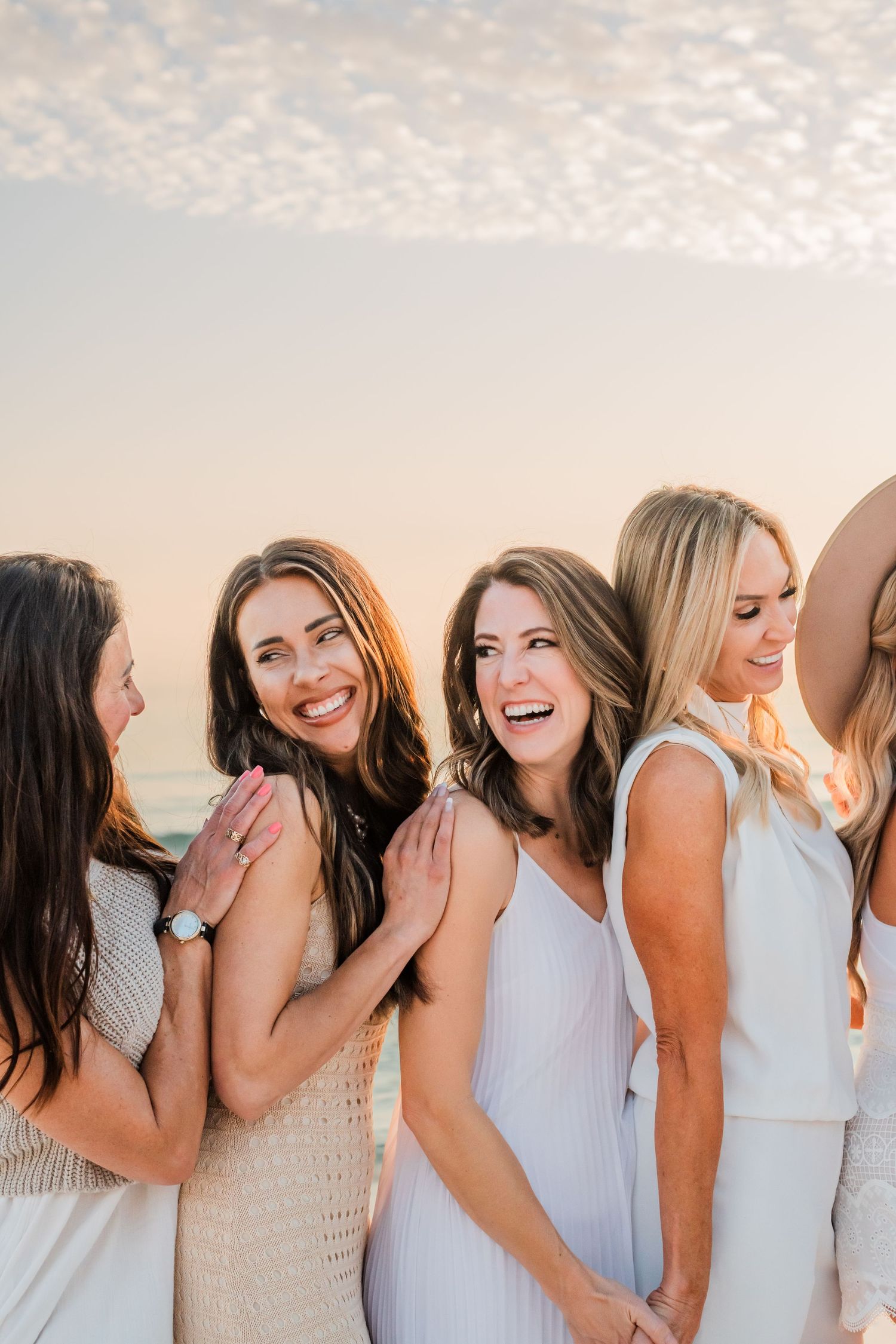 A group of friends laugh together while wearing light colored dresses during golden hour.