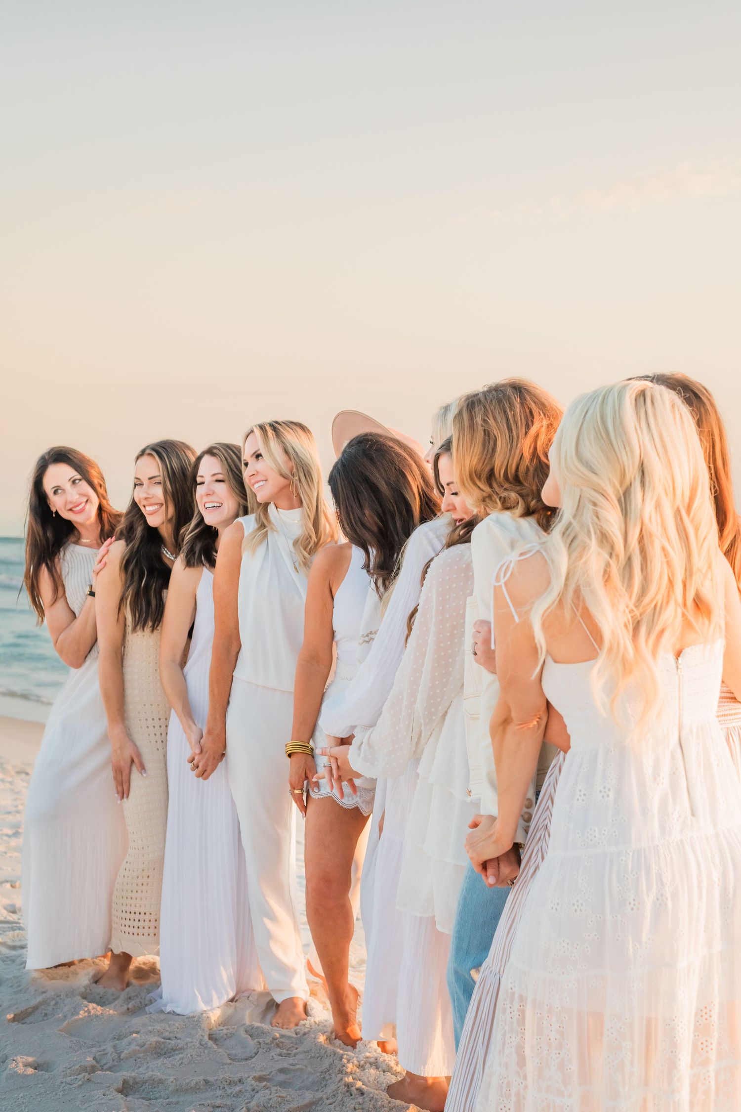 A group in white dresses stands together laughing and smiling on a beach at sunset.