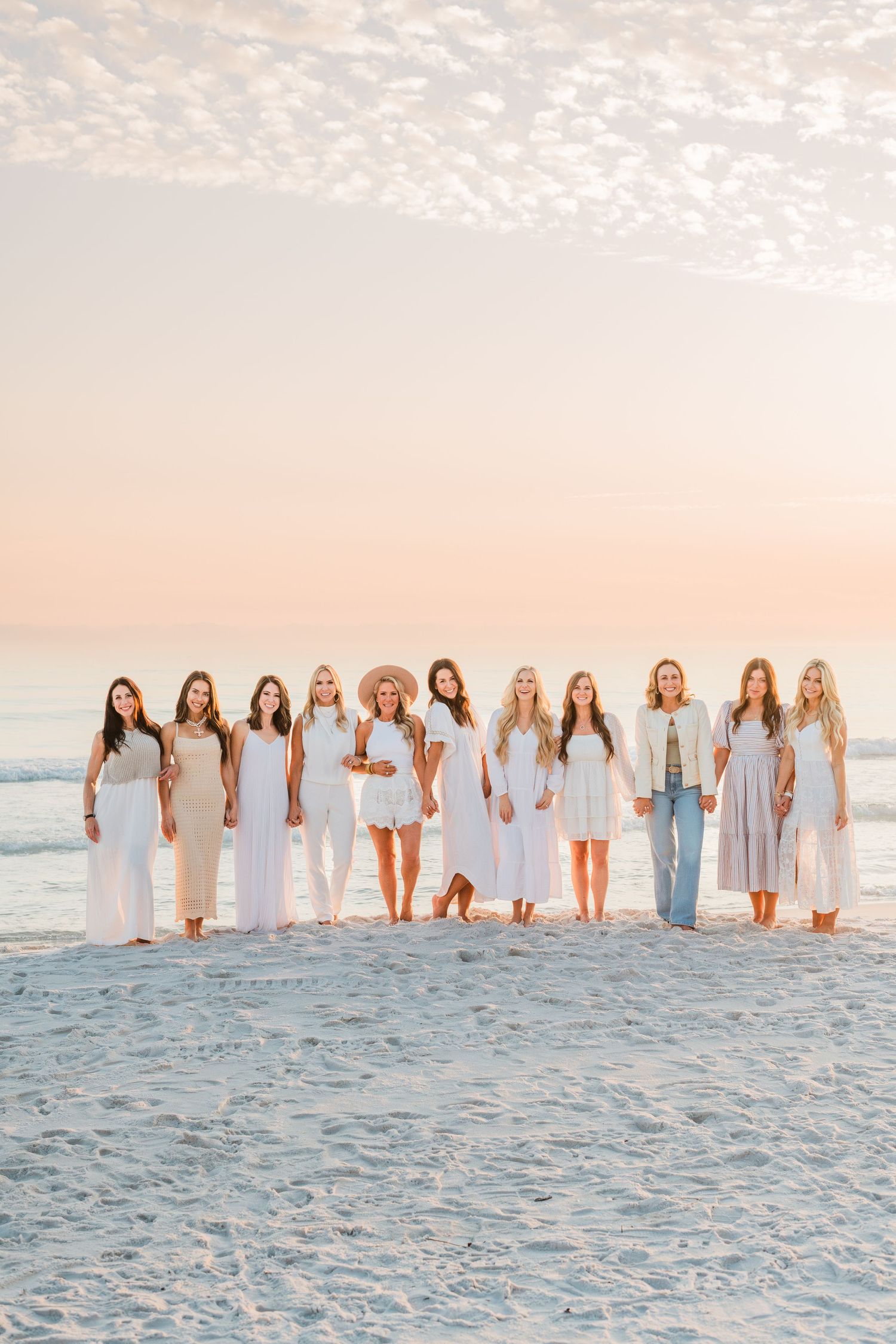 Group stands together on white sand beach at sunset wearing coordinated light colored dresses and outfits.
