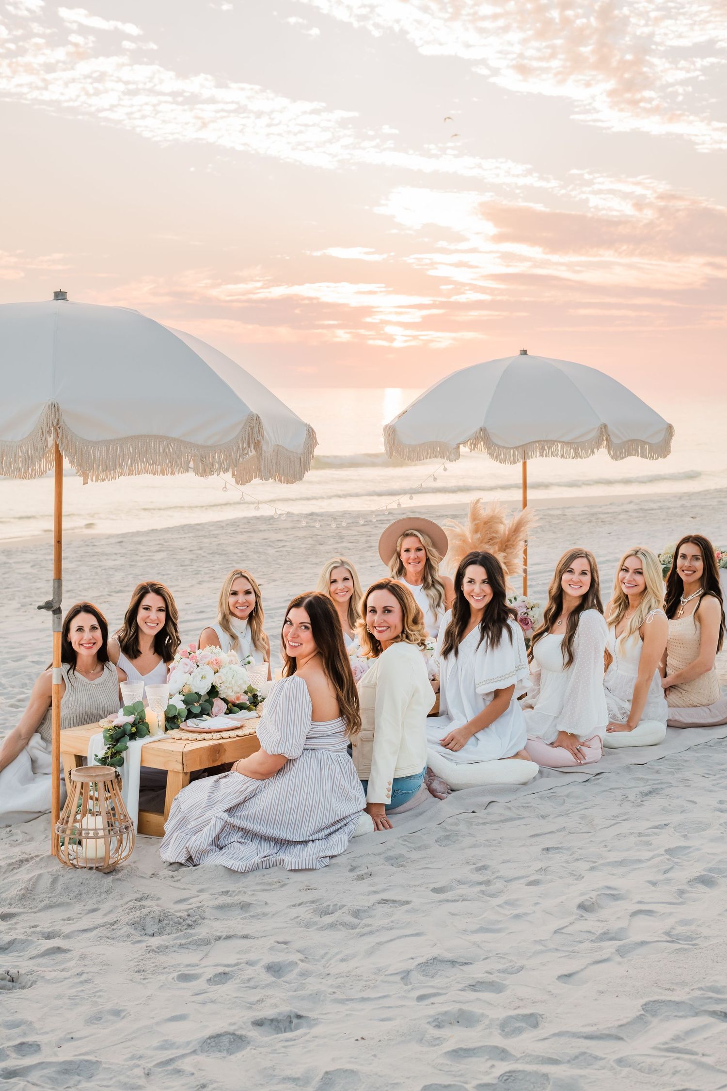 Friends in white and neutral dresses gather on a beach at sunset with umbrellas and decorative setup.