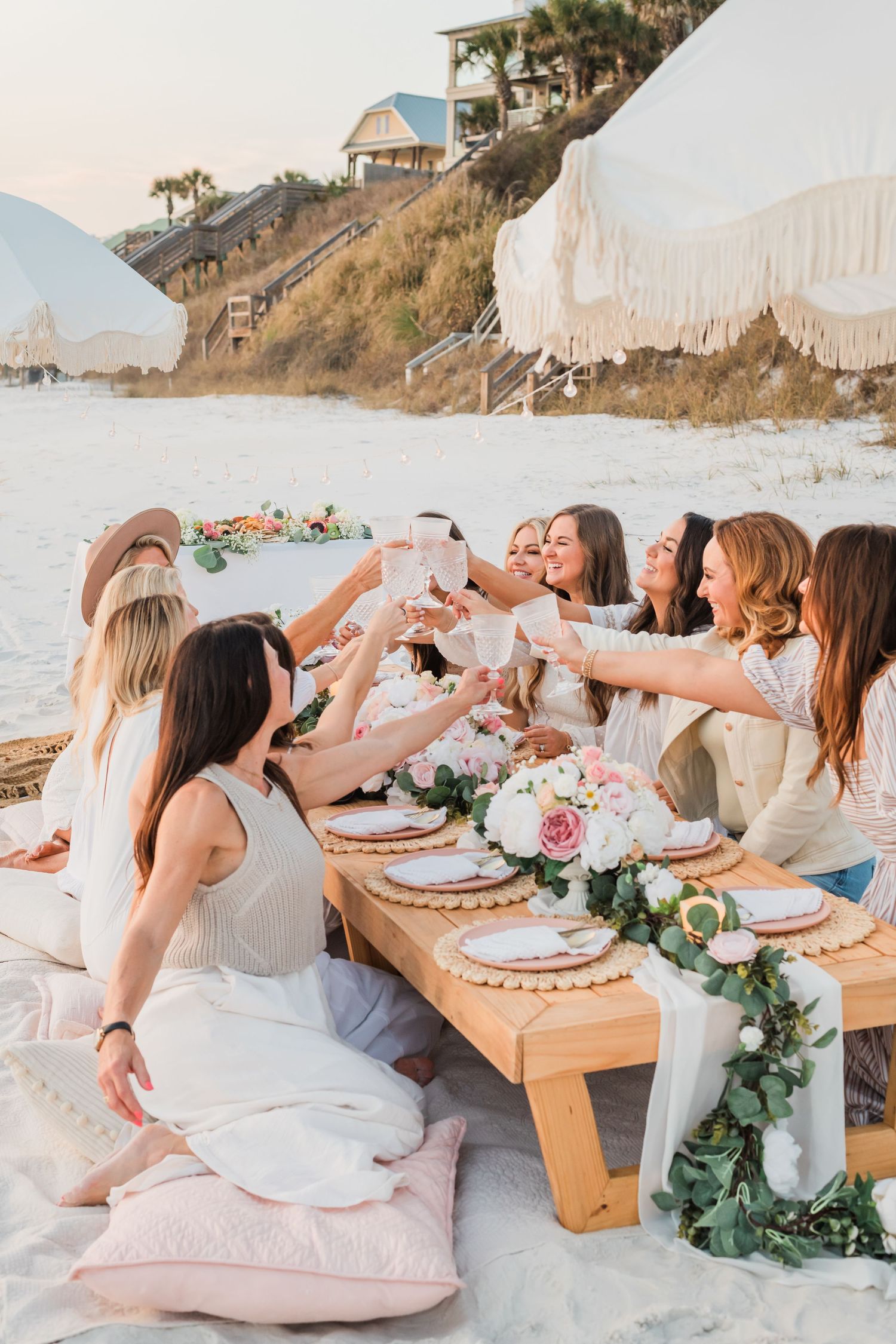 Beachside dinner party with friends toasting at sunset on a decorated wooden table with flowers and pillows on the sand.