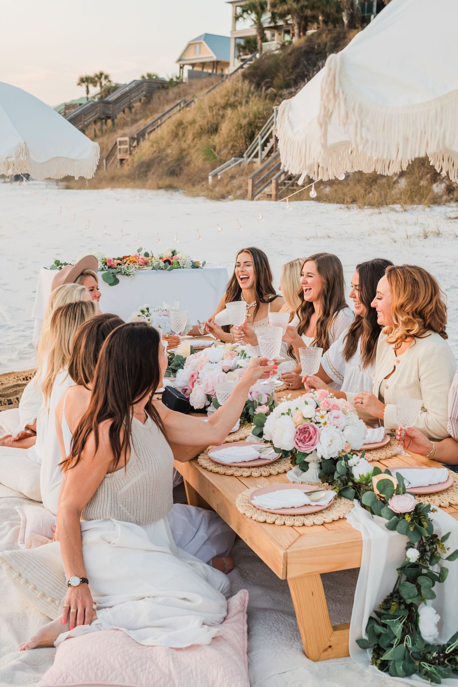 A beachside gathering with people in white attire enjoying food and drinks at a decorated low wooden table at sunset.