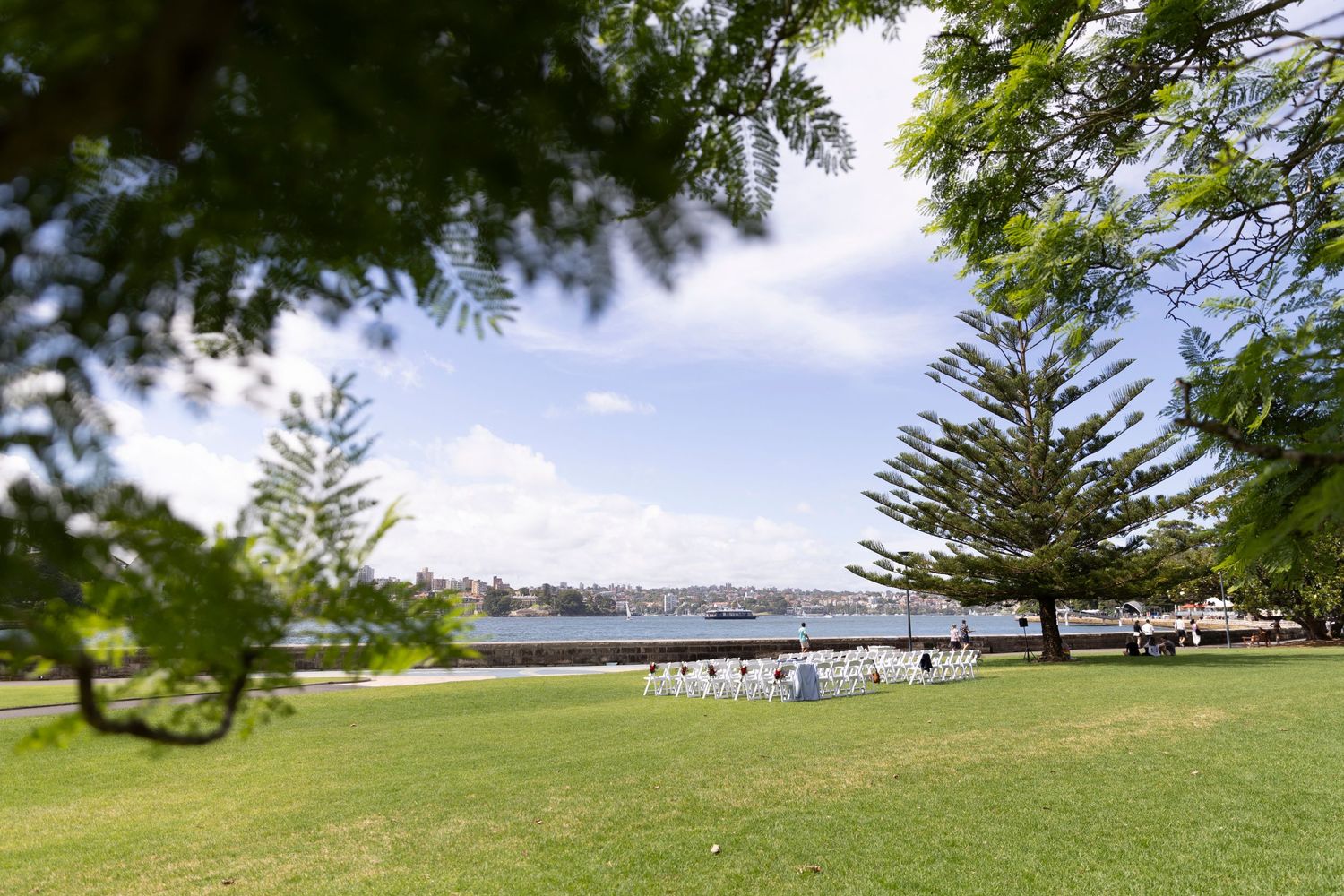 A scenic waterfront park setting with green lawn, pine trees and white chairs set up for an event.