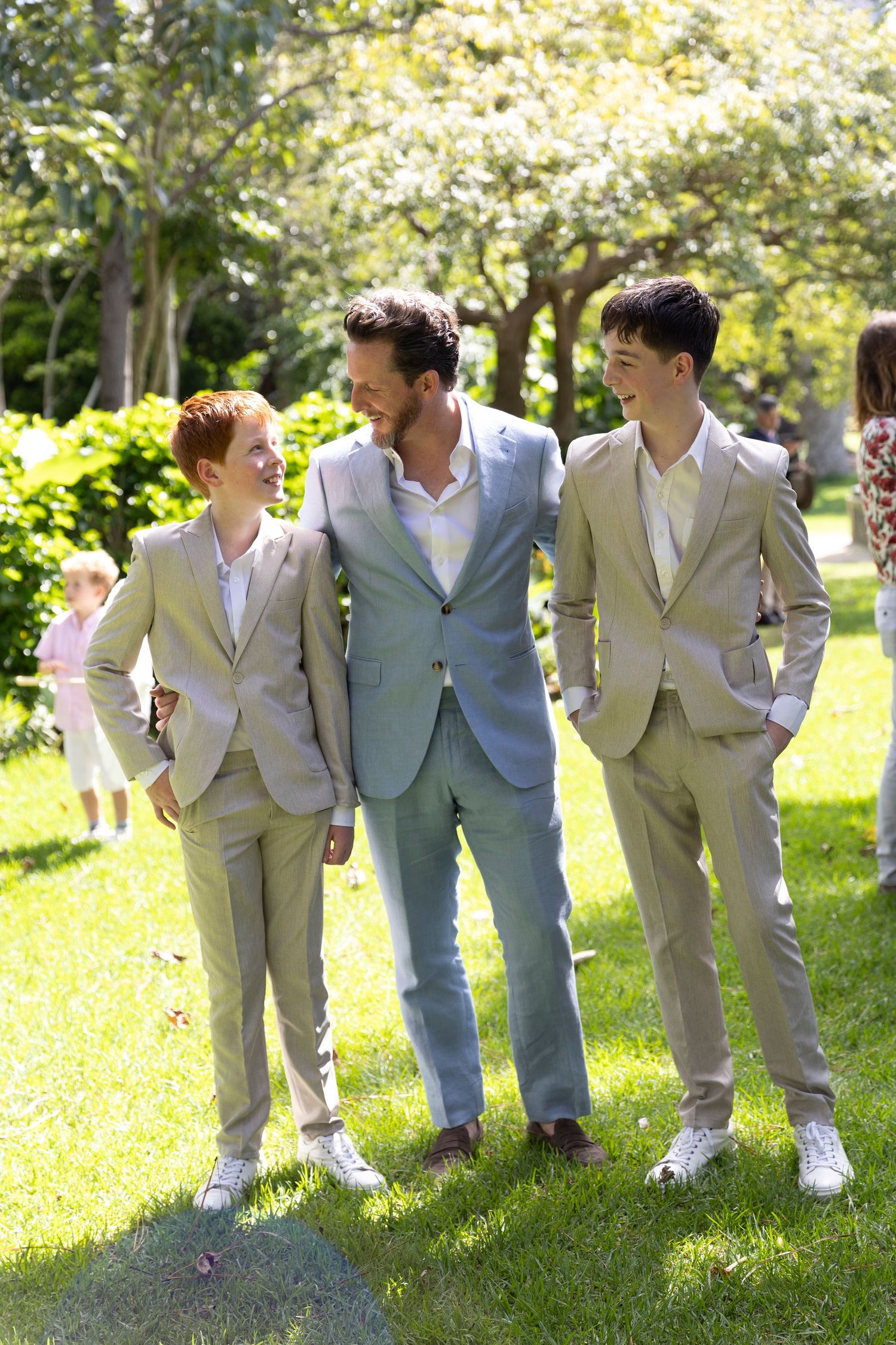 Three people in coordinating light-colored suits share a moment on sunny garden grounds.