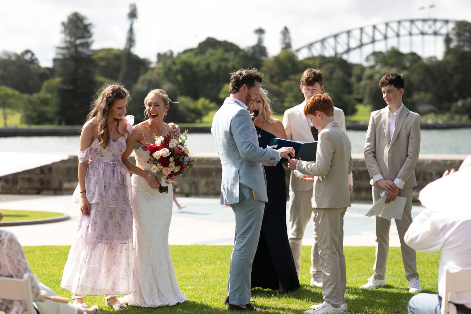An outdoor wedding ceremony by the water with attendants in pastel dresses and light colored suits.