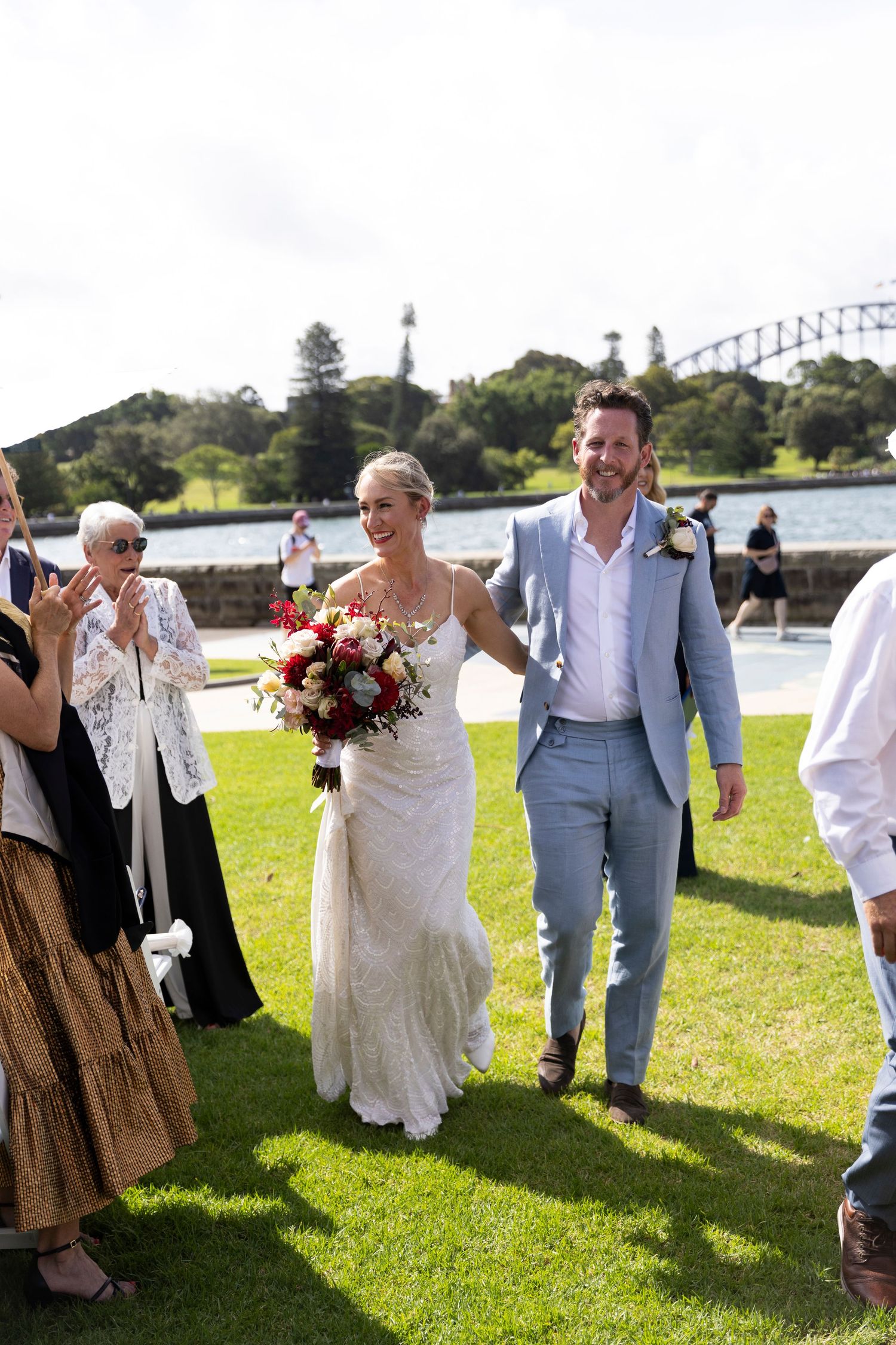 Wedding party celebrates on lawn with harbor bridge visible in background.