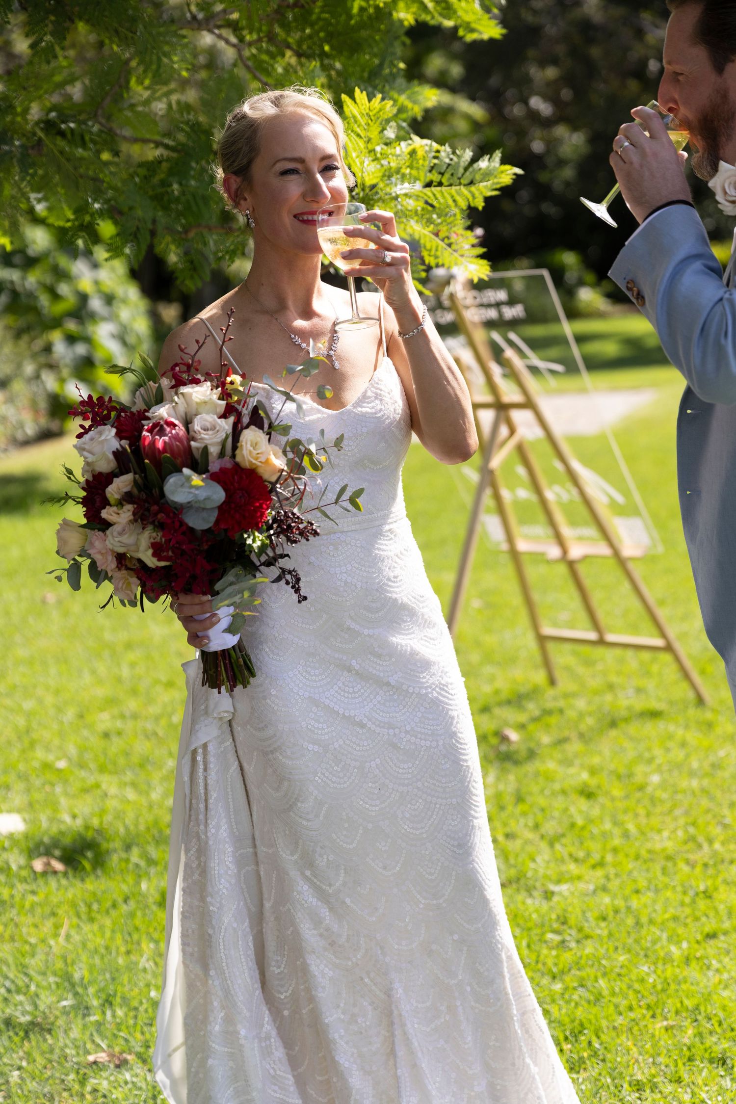 A bride in white holds a red bouquet while raising a glass during an outdoor celebration.