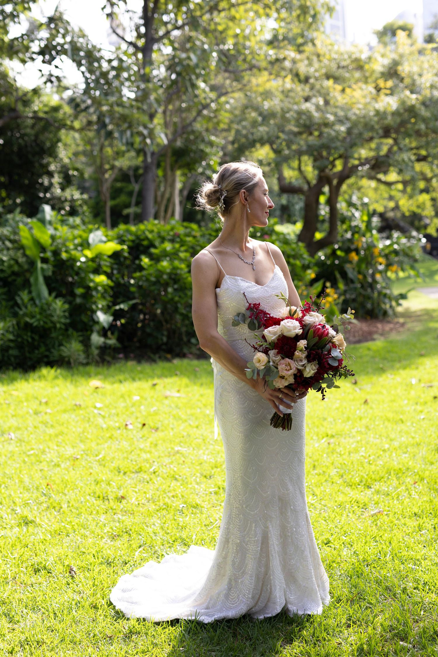 Bride in elegant lace gown poses with burgundy bouquet in sunny garden setting.
