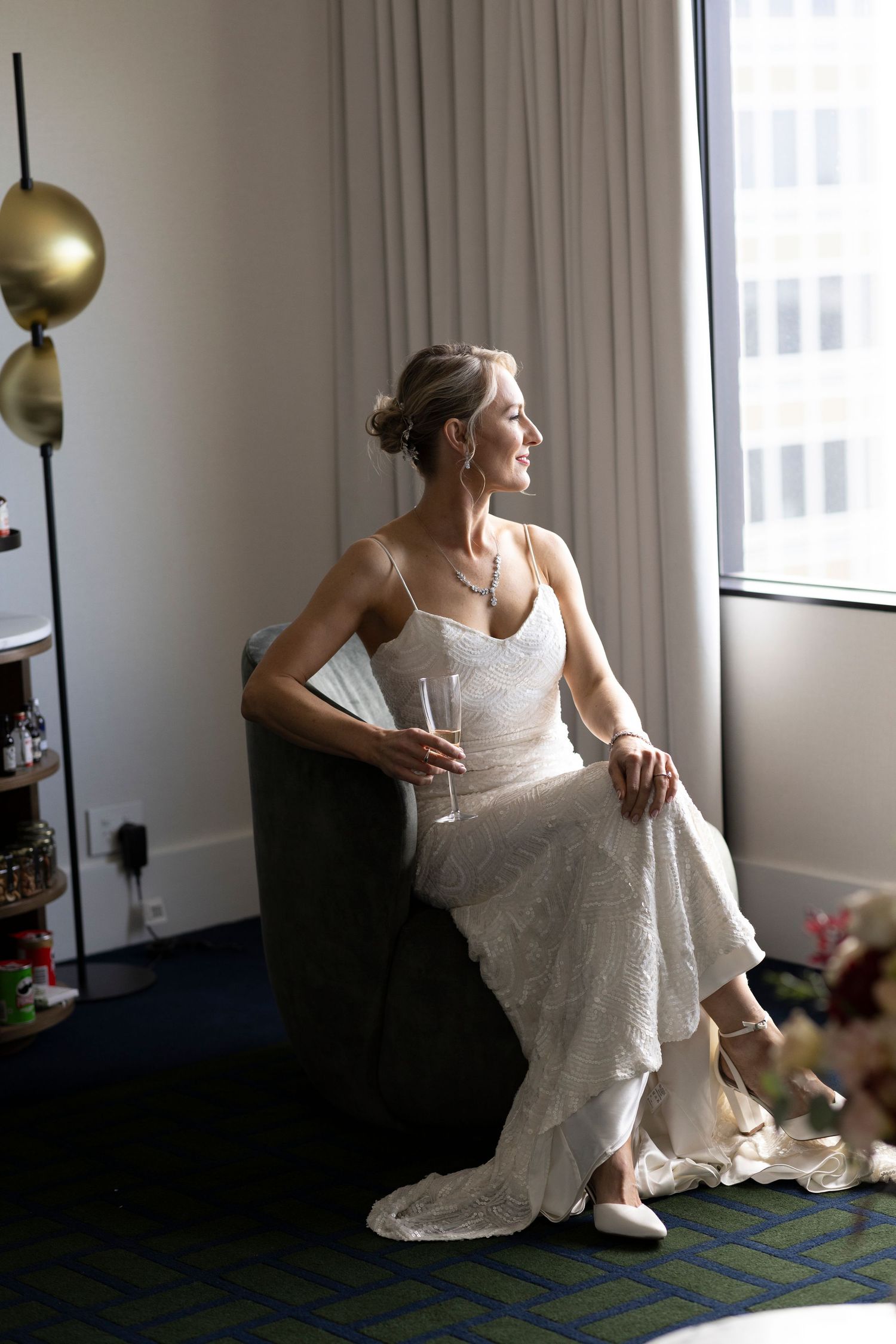 A bride in a lace wedding dress sits elegantly in a chair by a window with city views.