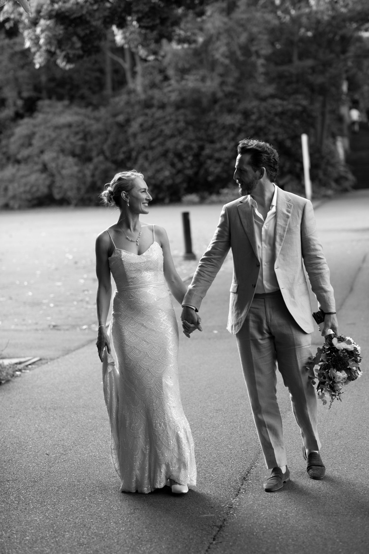 Black and white photo of wedding couple walking hand in hand on park path.