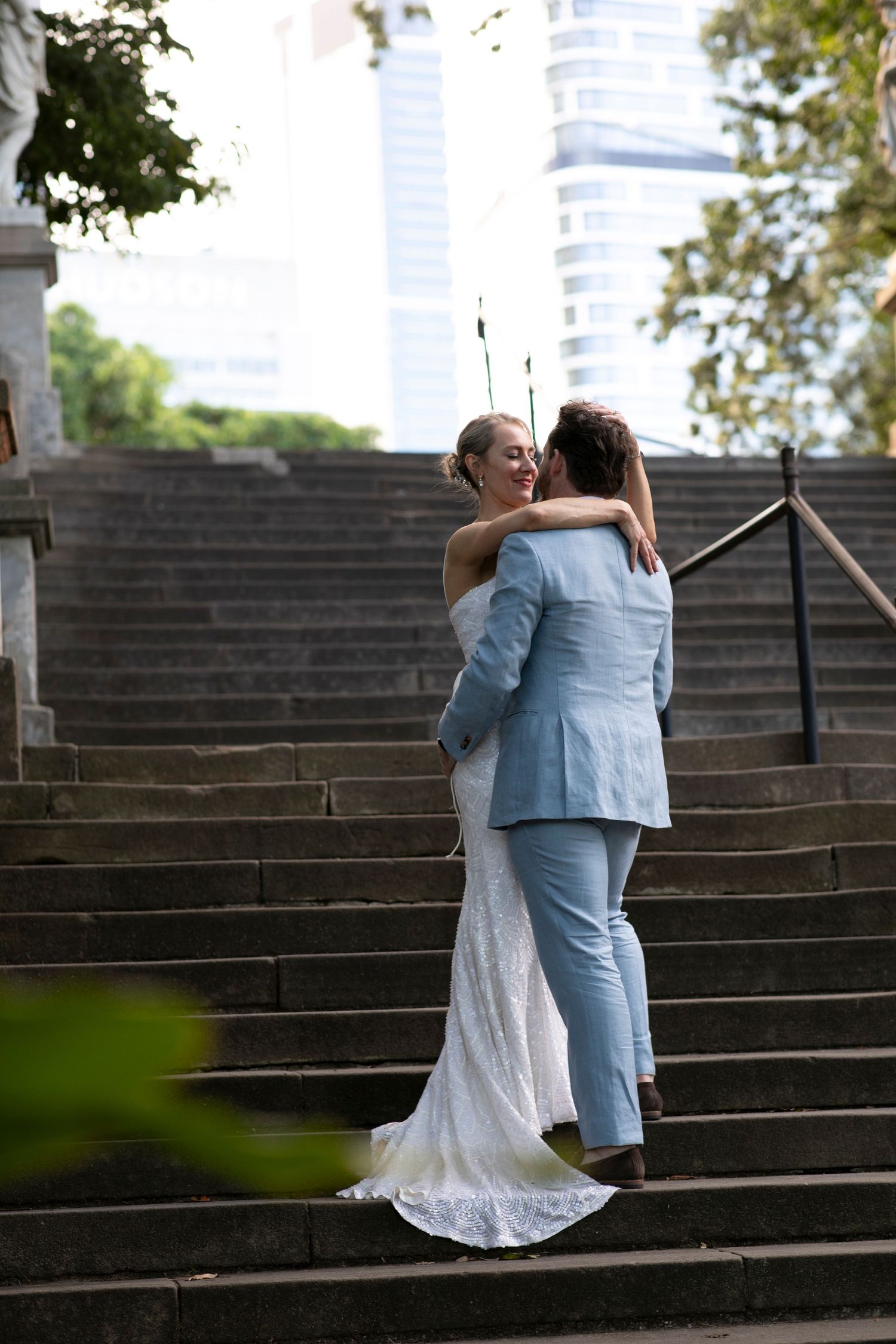 A couple embracing on grand stone steps with city buildings visible in the background.