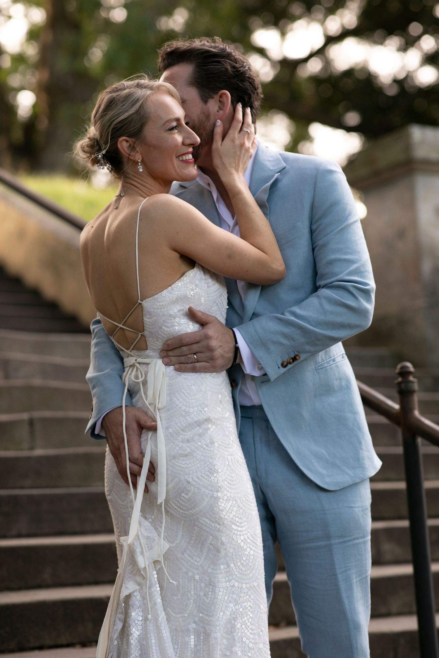 A romantic wedding moment on stone steps with the bride in white and groom in light blue embracing tenderly.
