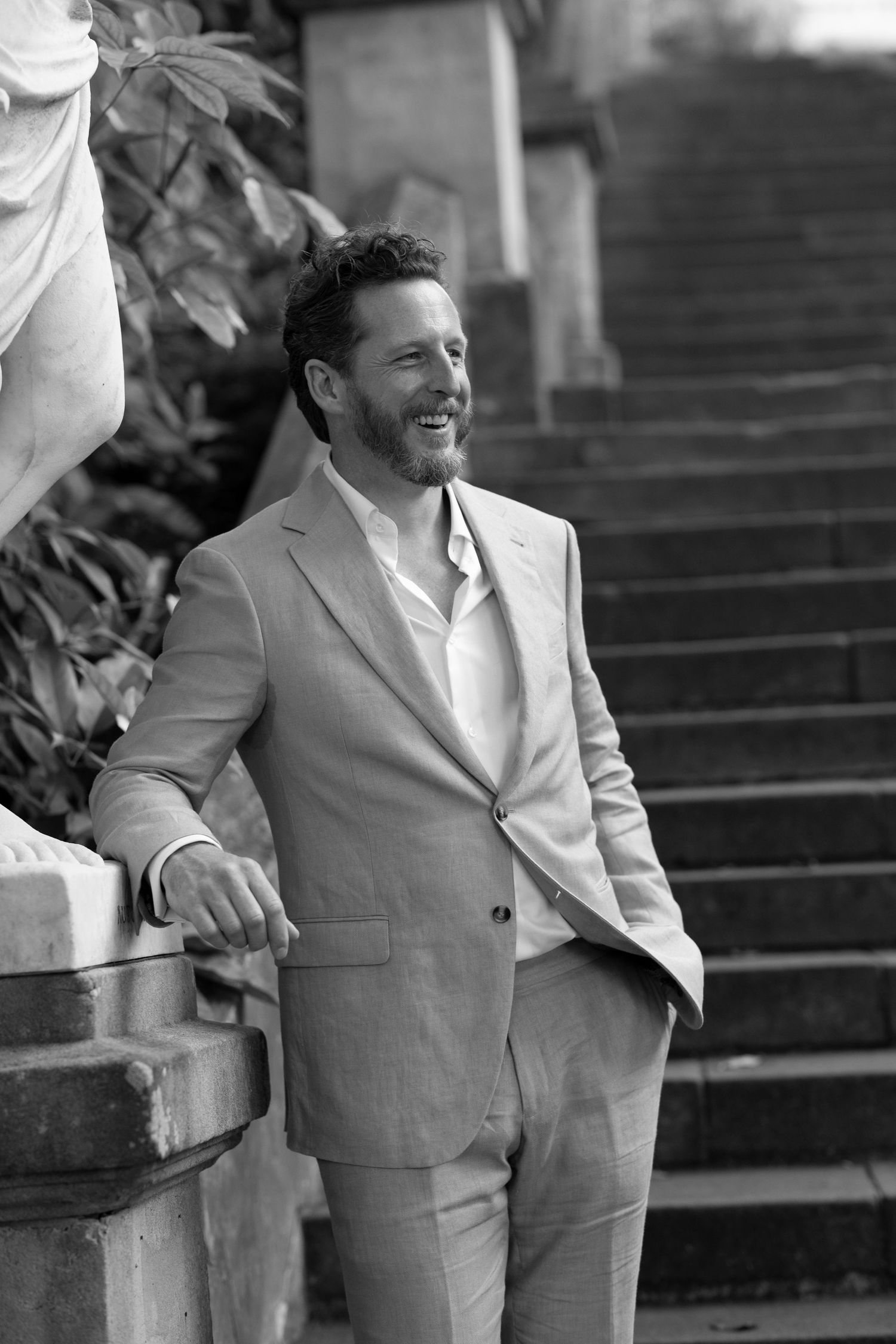 Man in light gray suit stands smiling on outdoor stone steps in black and white photo.