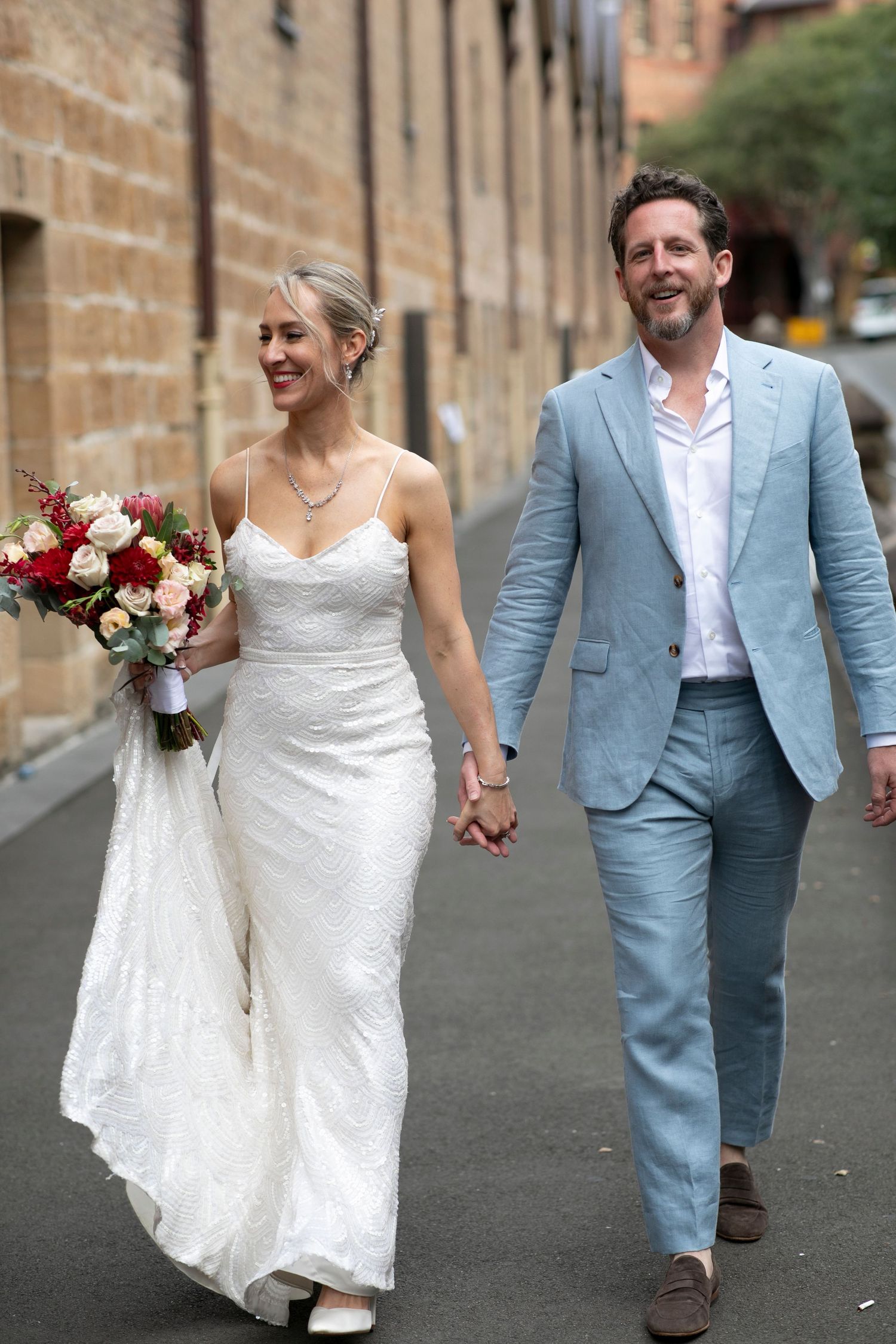 Wedding couple walking hand in hand on urban street with bride carrying red and white bouquet.