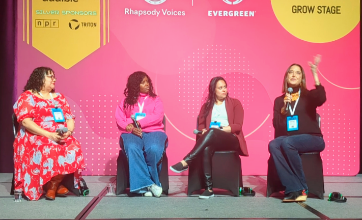 A panel discussion featuring four speakers, Monika Aldarondo is speaking, they are seated on a stage with a vibrant pink backdrop.