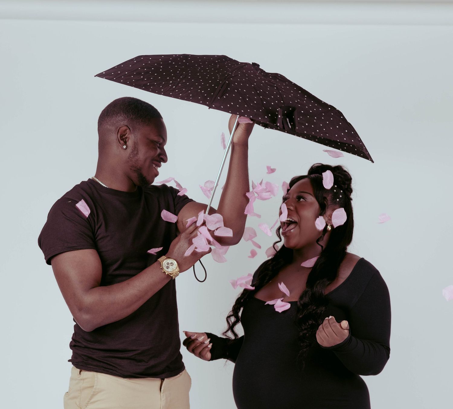 Couple sharing a joyful moment under a polka dot umbrella with pink flower petals falling around them.