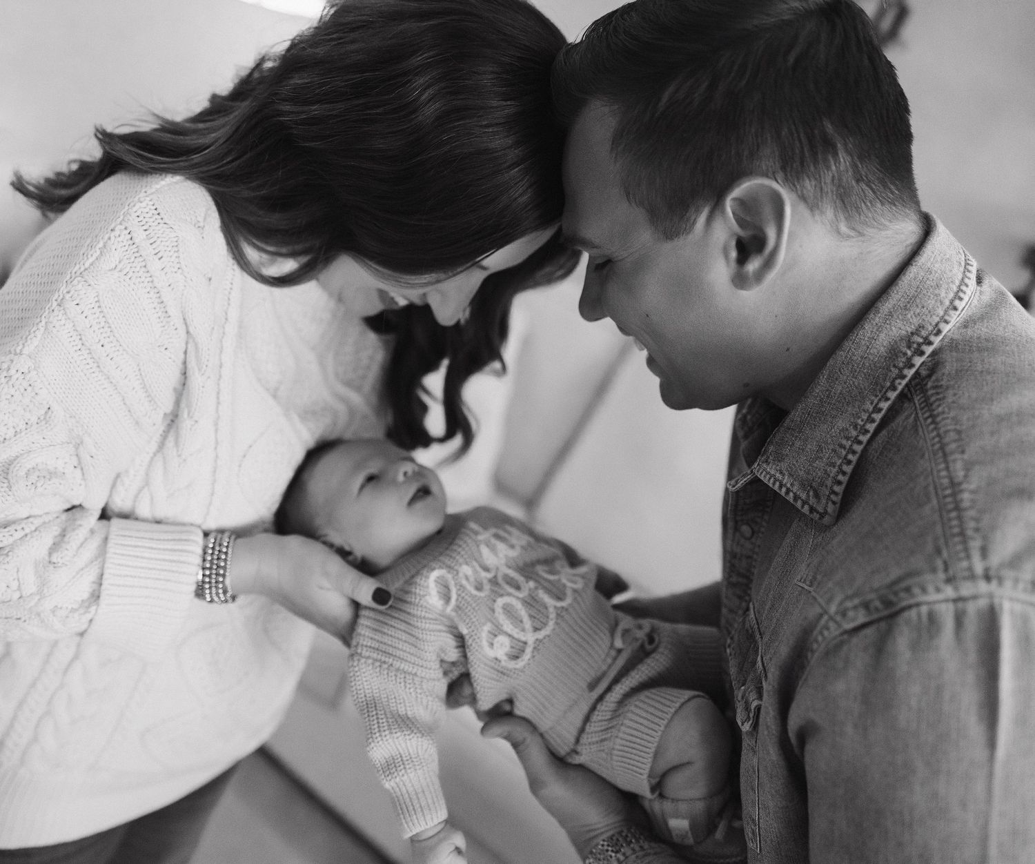 A tender black and white photo of parents lovingly looking down at their baby in an intimate family moment.