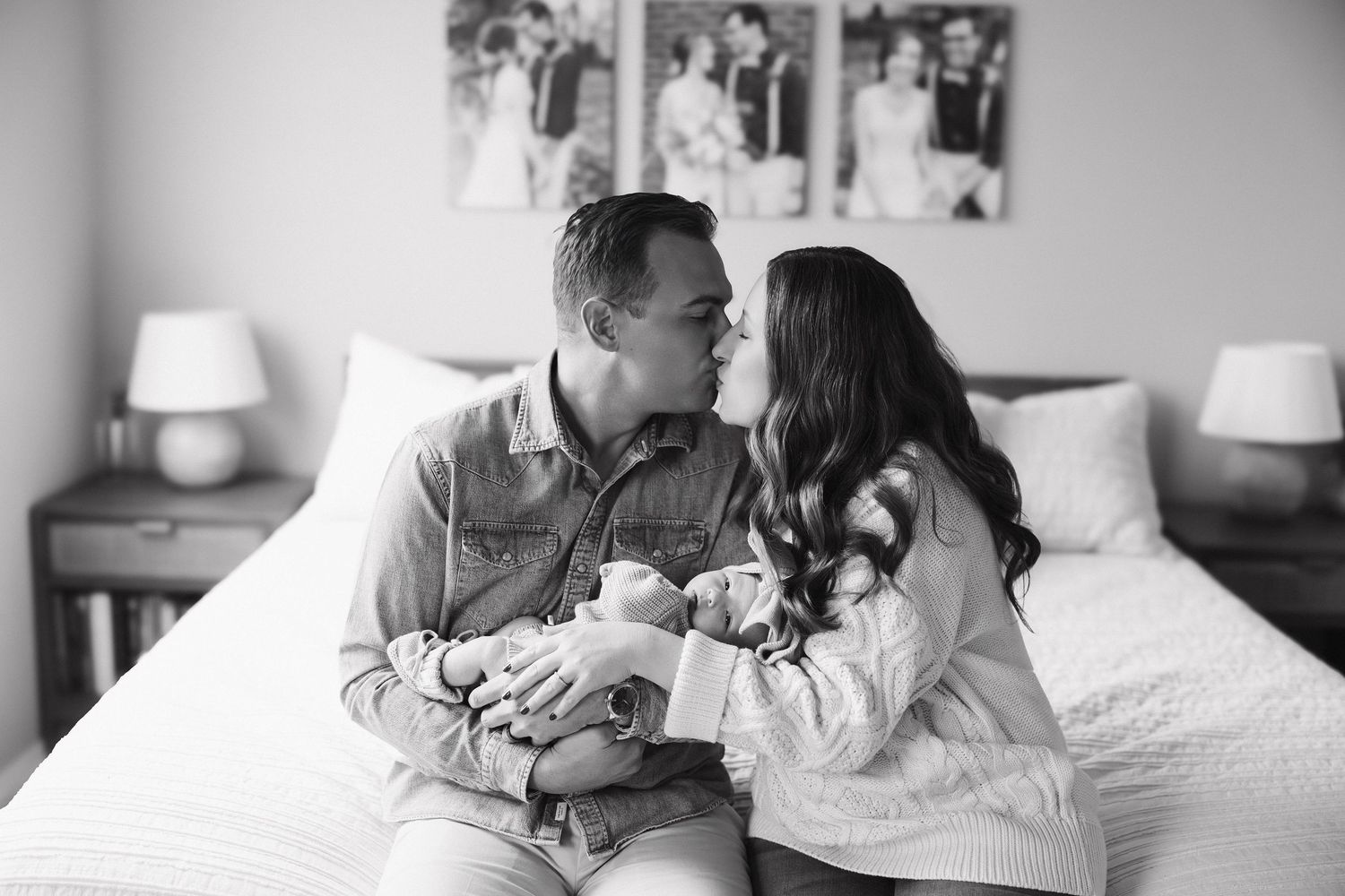 A tender black and white moment between a couple sitting on a bed with framed photos on the wall behind them.