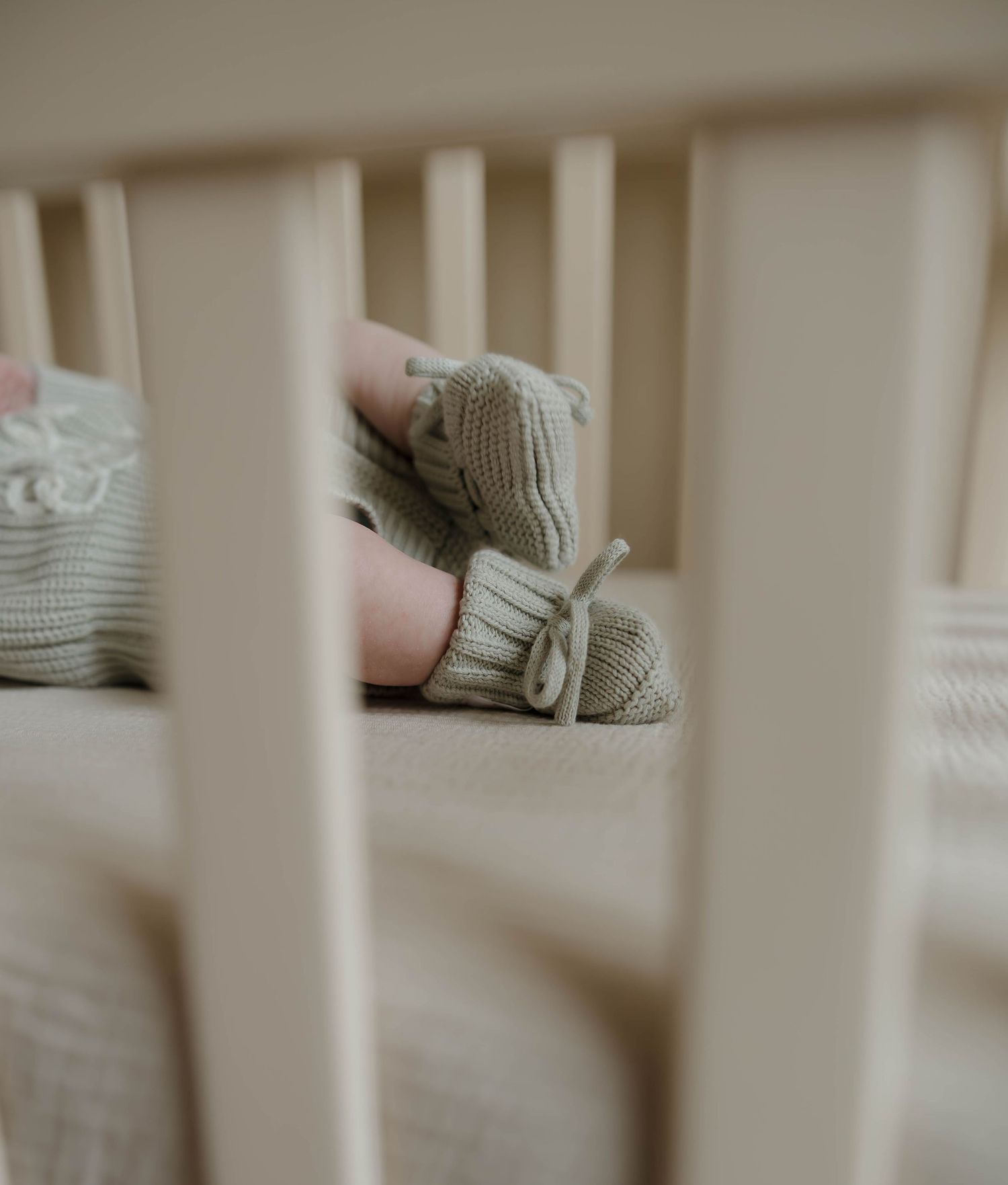 White crib bars frame a gentle scene of parent cradling infant in muted, neutral tones.