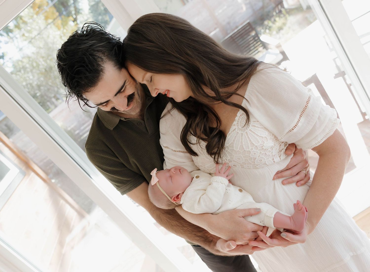 A couple in casual attire lovingly holds their newborn baby near a bright window in a tender family moment.
