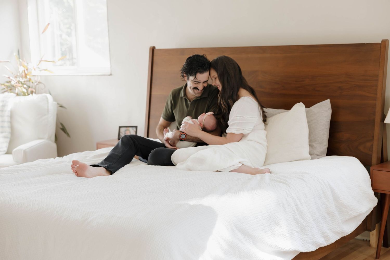 A sequence of intimate family moments on a white bed with wooden headboard in a bright bedroom.