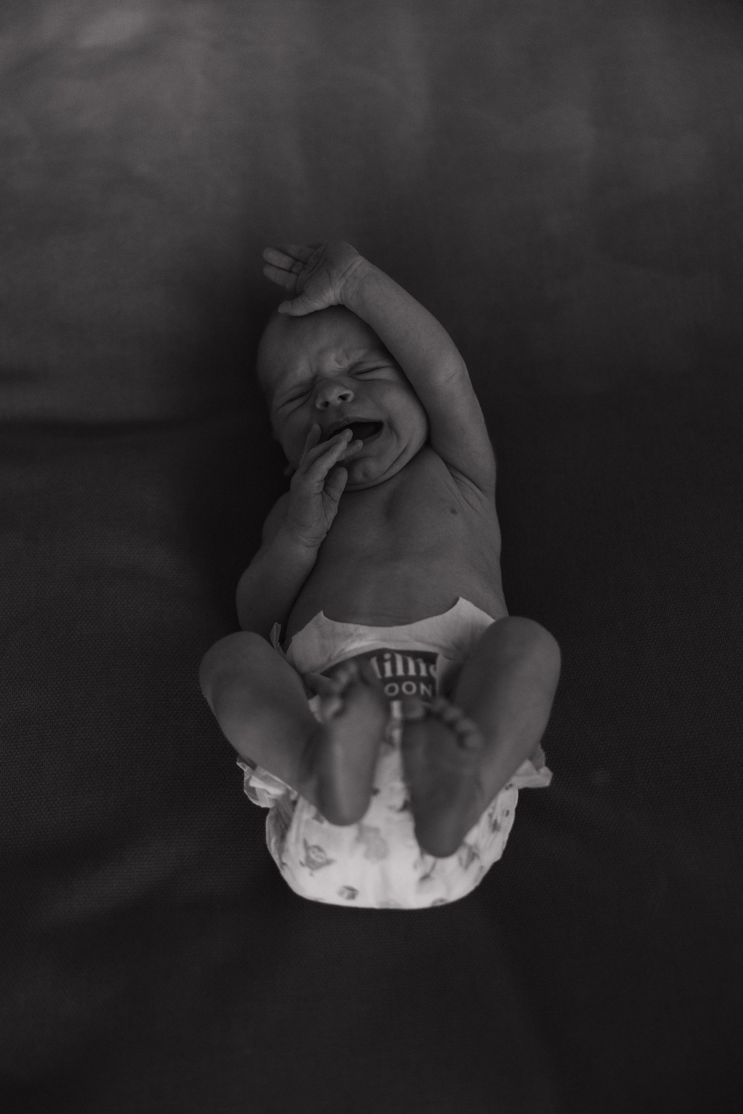 Black and white overhead view of a newborn baby lying on a dark background showing full body perspective.