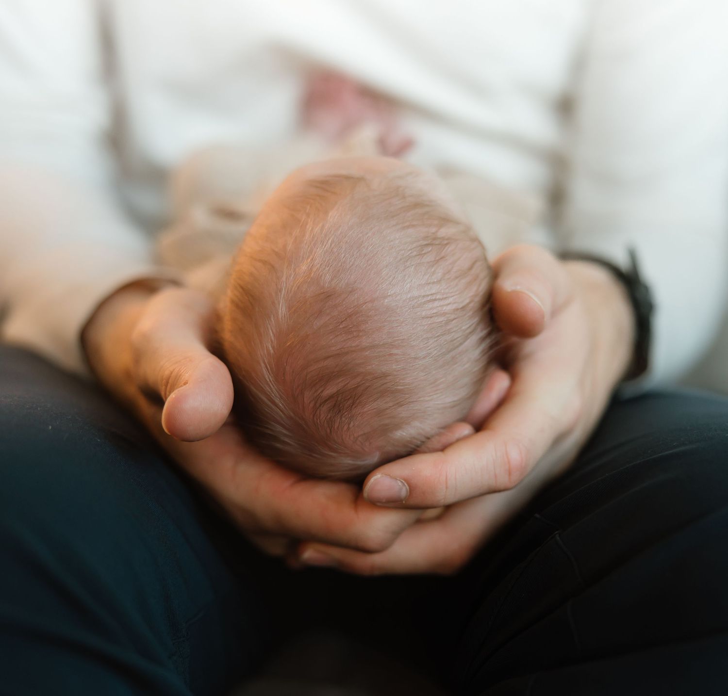 Gentle hands cradle a newborn baby's head with soft reddish hair against a light background.