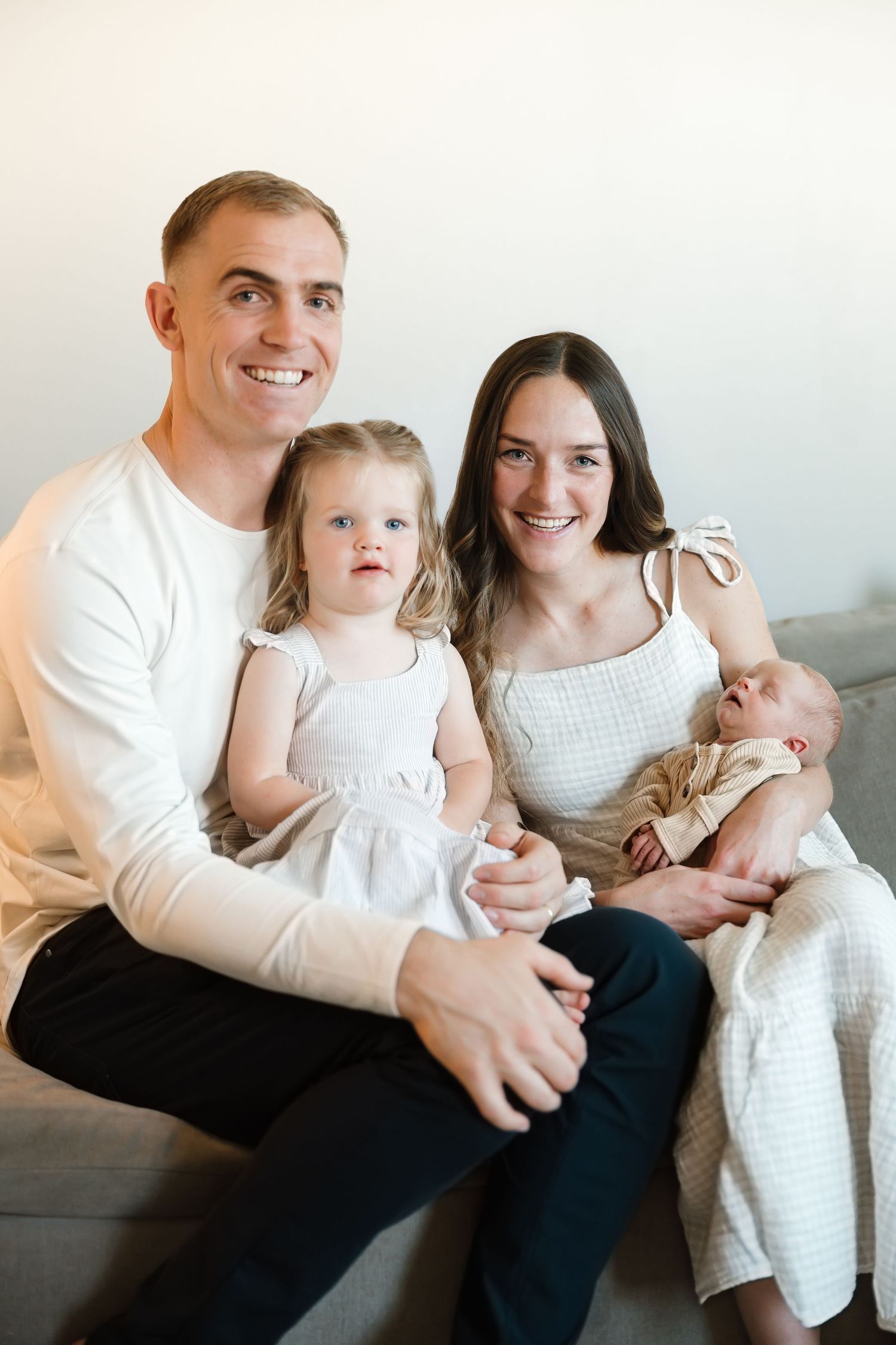 A family sits together on a gray couch wearing coordinated white and neutral outfits for a casual portrait session.