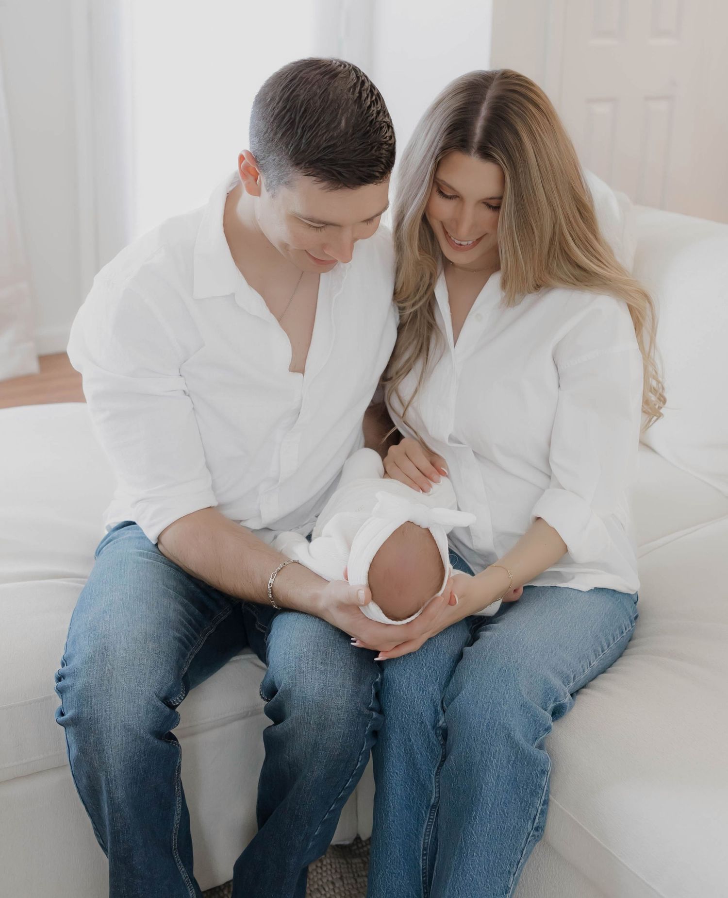A couple in white shirts and jeans sits together on a white couch in a bright, minimalist setting.