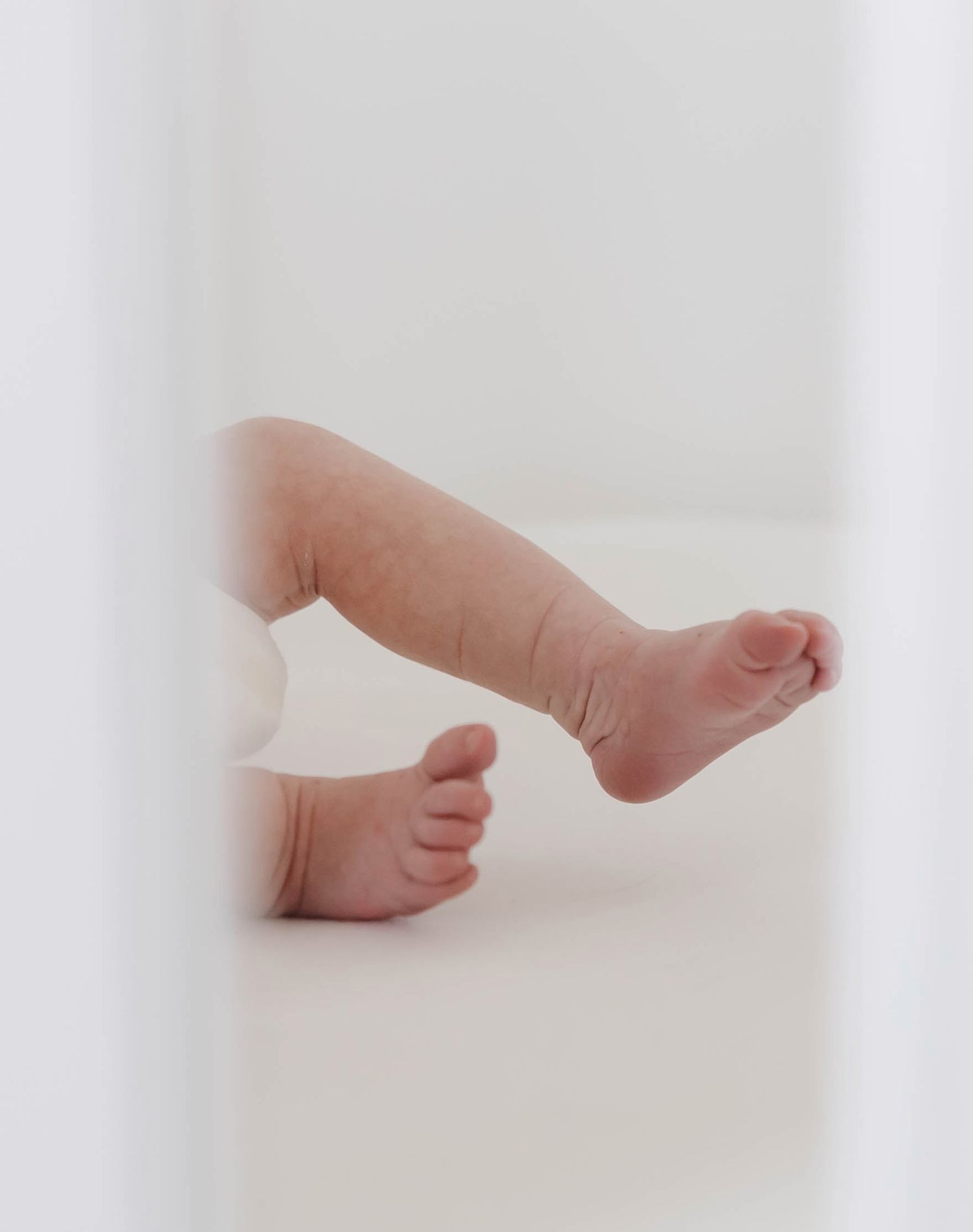 Tiny baby feet peek out from behind a white surface in a soft, minimalist newborn photograph.
