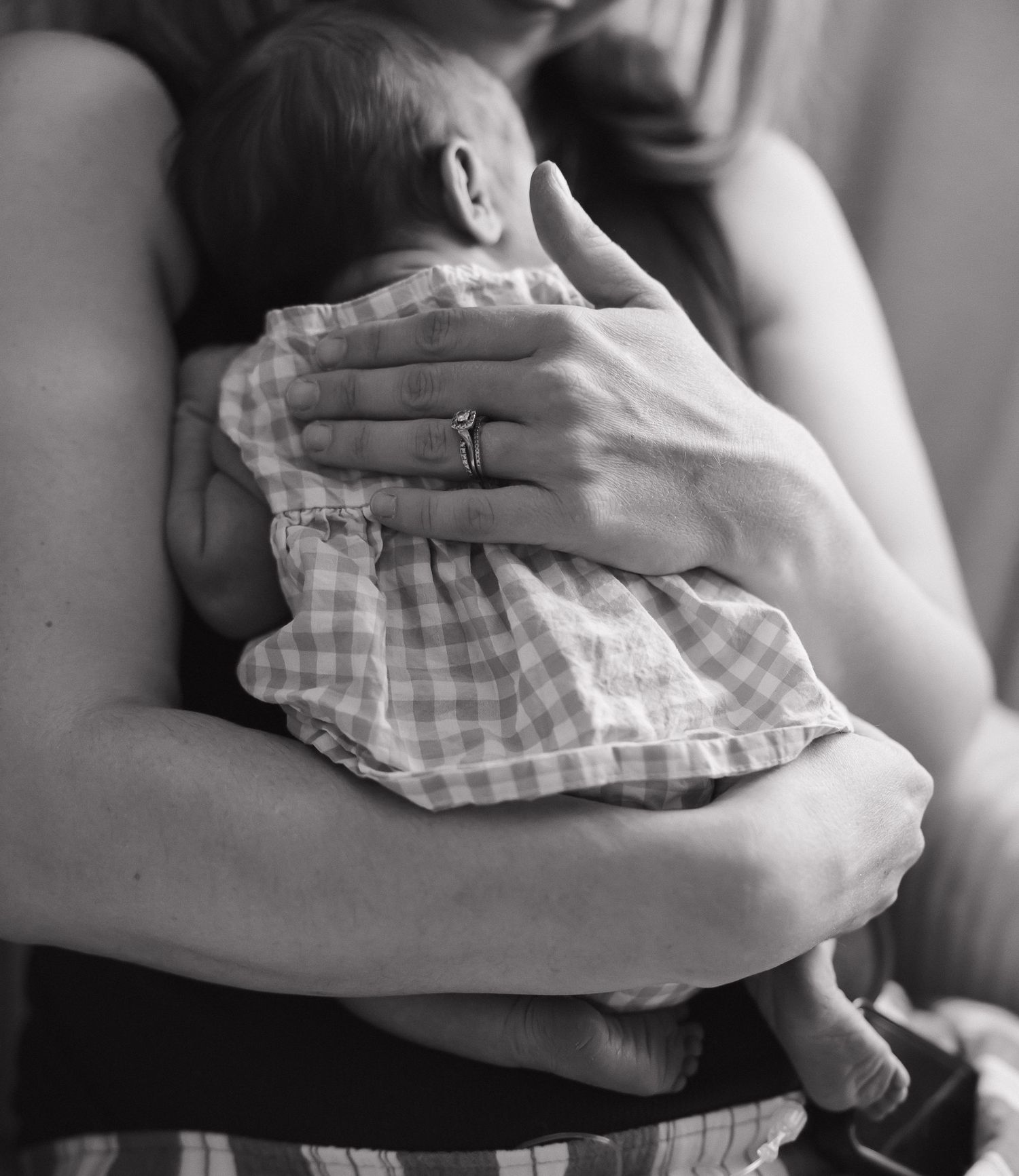Black and white image shows hands holding a small child wearing a checkered dress.