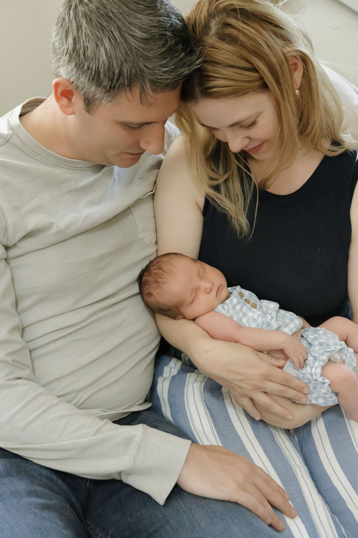 A couple sits together on a couch holding and gazing lovingly at their newborn baby.