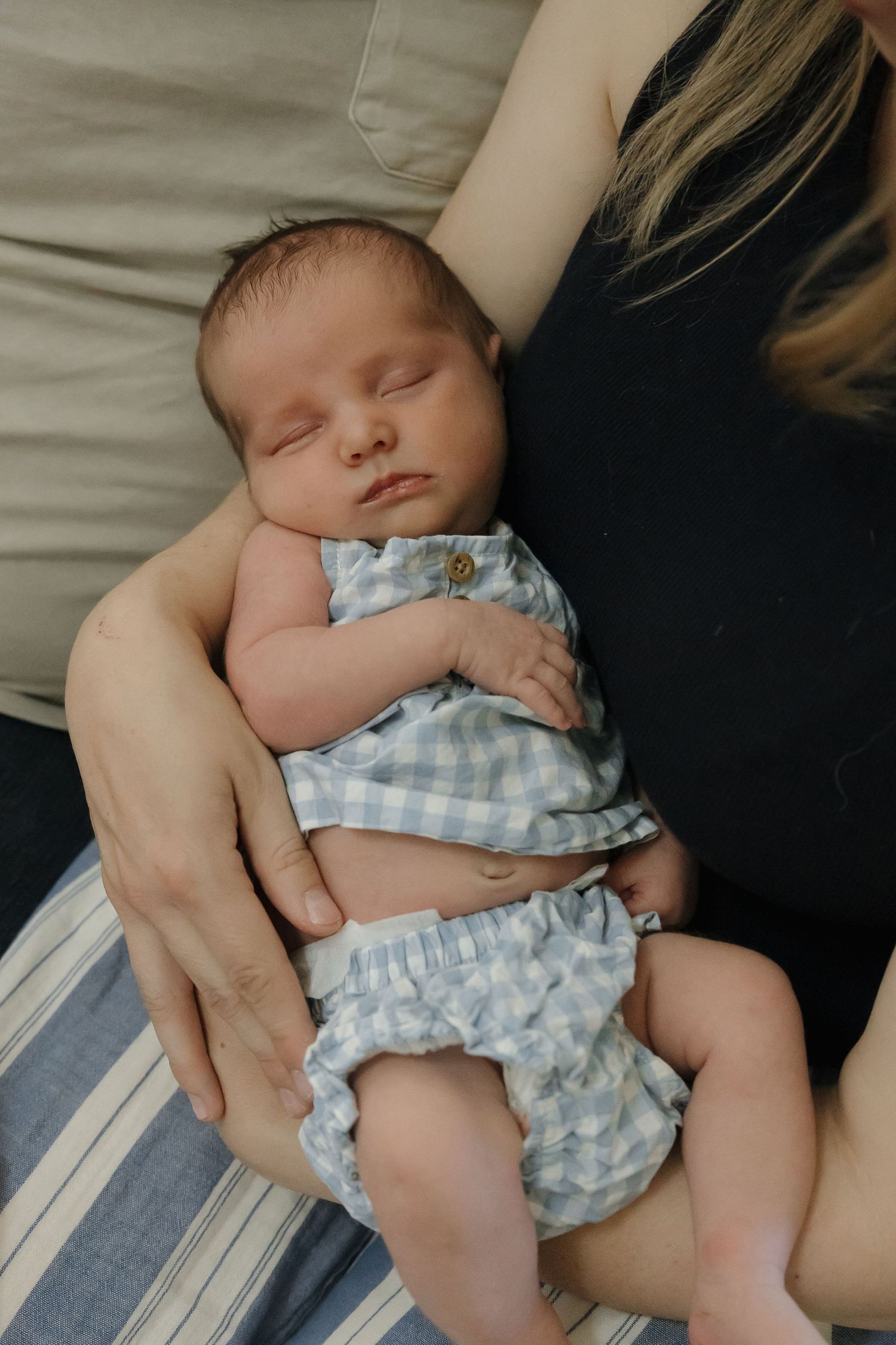 A sleeping baby wearing a blue gingham outfit rests peacefully in someone's arms.