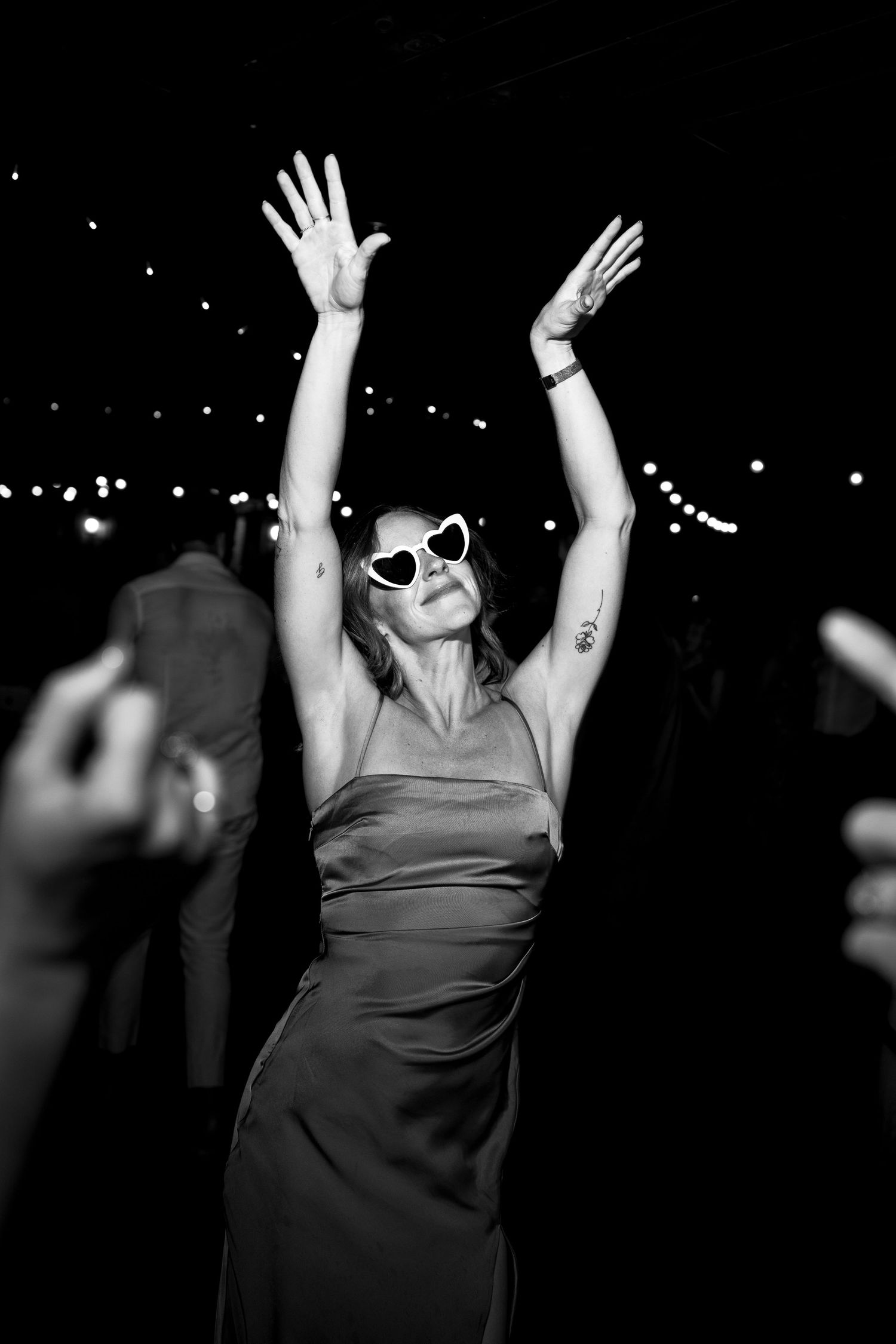 Woman in formal dress and sunglasses dancing with arms raised under string lights.